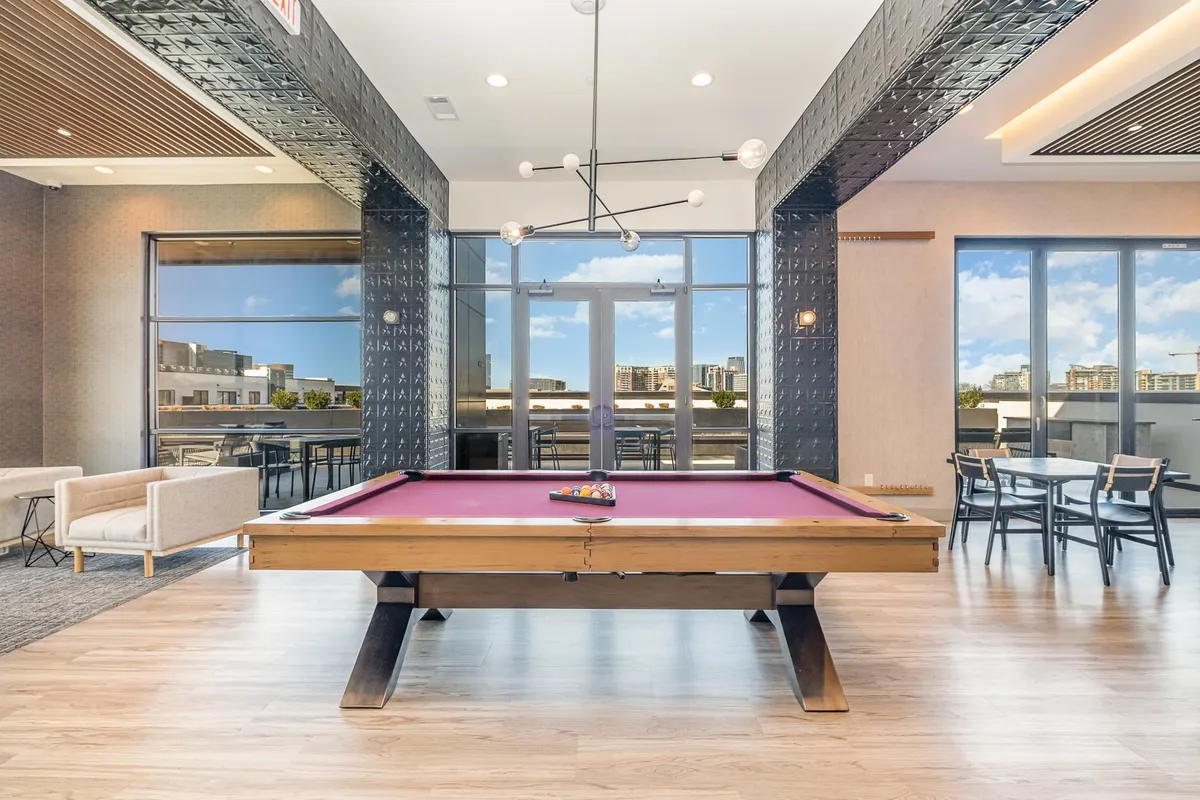 A lively, modern rooftop lounge interior with a pool table and large windows showcasing city views. The space is designed for social gatherings and relaxation, emphasizing both entertainment and comfort.