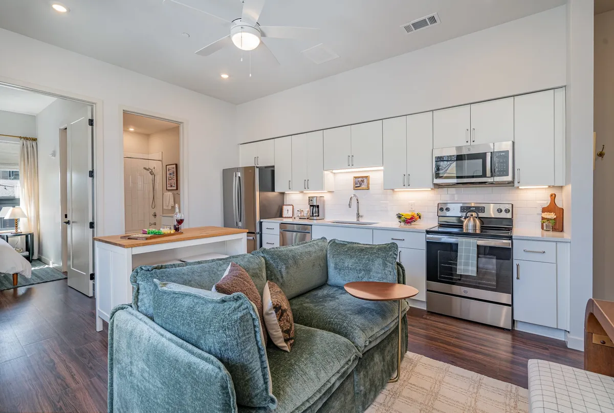 An open-plan living space with a comfortable sofa, central kitchen island, gleaming kitchen appliances, and an open doorway leading to what appears to be a private bedroom. The setup demonstrates the fluid transition between shared and private areas.