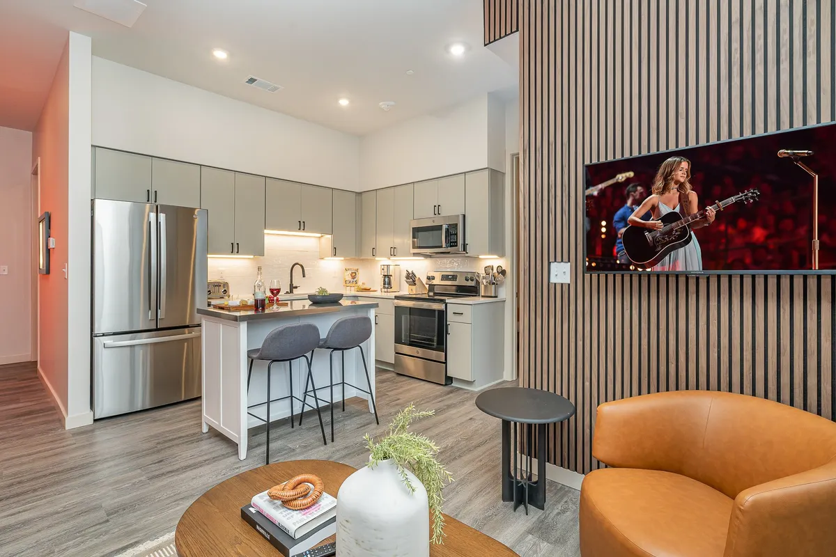 A bright, contemporary open-plan kitchen and living area featuring a spacious kitchen island with seating, modern appliances, stylish wall art, and a cozy seating area perfect for gathering. The TV is displaying a live concert, suggesting a vibrant, music-themed atmosphere ideal for entertaining or relaxing with friends and family.