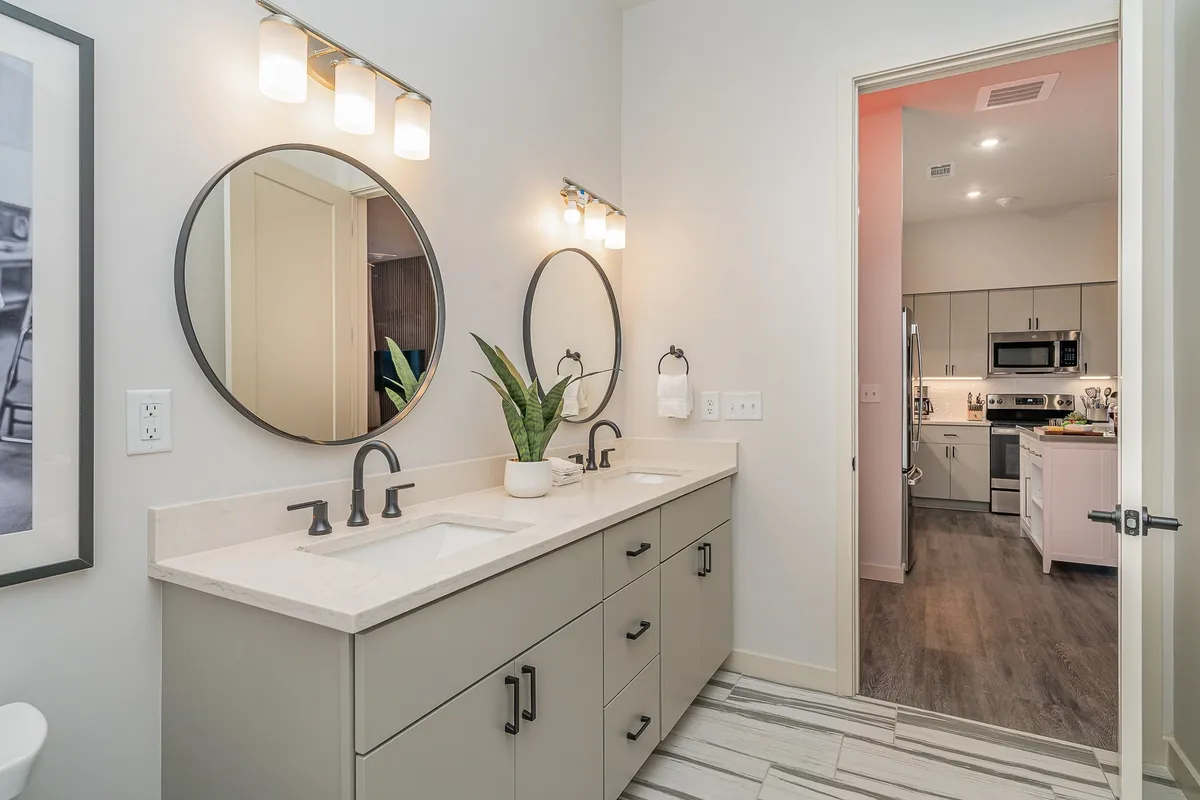 A modern bathroom featuring a double sink vanity, symmetrical mirrors, elegant lighting fixtures, and an indoor plant adding a fresh touch. The space is bright, organized, and inviting, perfect for streamlined morning routines or unwinding after a day out.