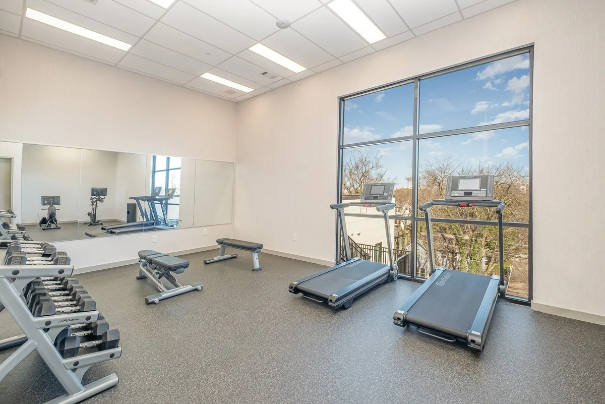 A bright, modern gym featuring treadmills, weights, large windows offering natural light, and mirrored walls. The space is designed for both cardio and strength training, with ample room for movement and social interaction.