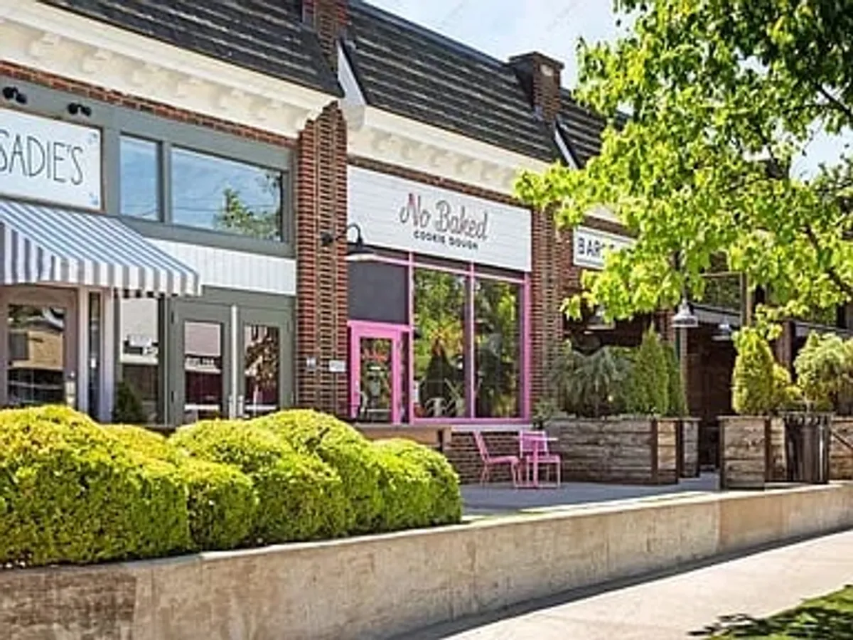 This cheerful exterior shot captures the storefront of No Baked cookie shop, flanked by green shrubs and featuring an outdoor seating area. The setting is bright and inviting, ideal for family outings to local treat destinations.