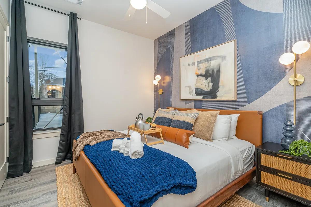 A serene bedroom scene with a well-made bed topped with a blue throw blanket, large artwork above the headboard, modern light fixtures, and curtains gently framing a sunlit window. The ambiance is calm and intimate, ideal for morning coffee or cozy evenings in.