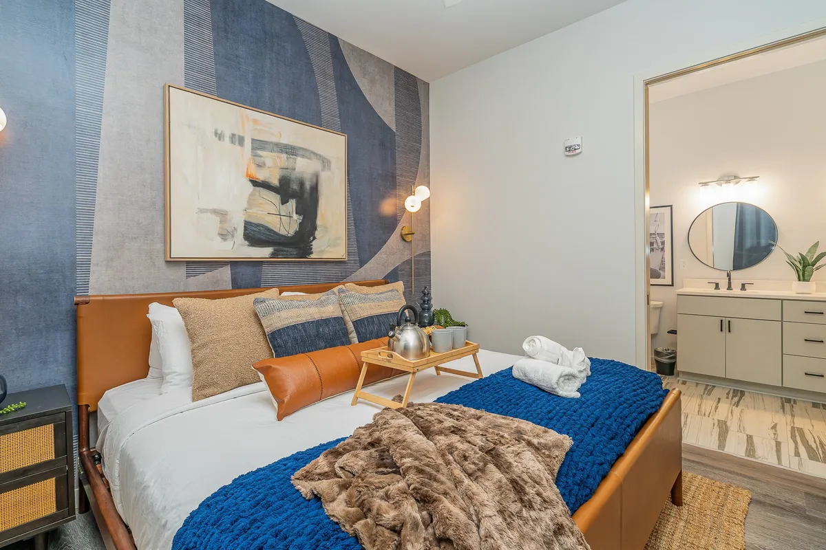 A bedroom featuring a king-size bed with a stylish leather headboard, textured blue throw blanket, abstract wall art, decorative pillows, and a nightstand with a lamp. The space is softly lit, exuding a sense of comfort and tranquility perfect for relaxation and unwinding after a busy day.