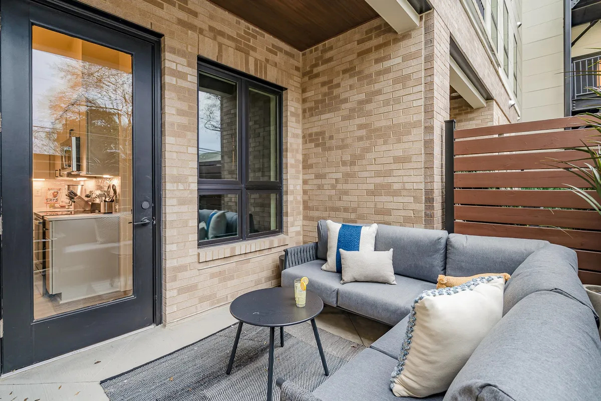 A cozy patio scene featuring a grey sectional sofa with decorative pillows, a round black table, and a sliding glass door opening to a private outdoor space. The mood is intimate and inviting, suitable for enjoying drinks and quiet moments.