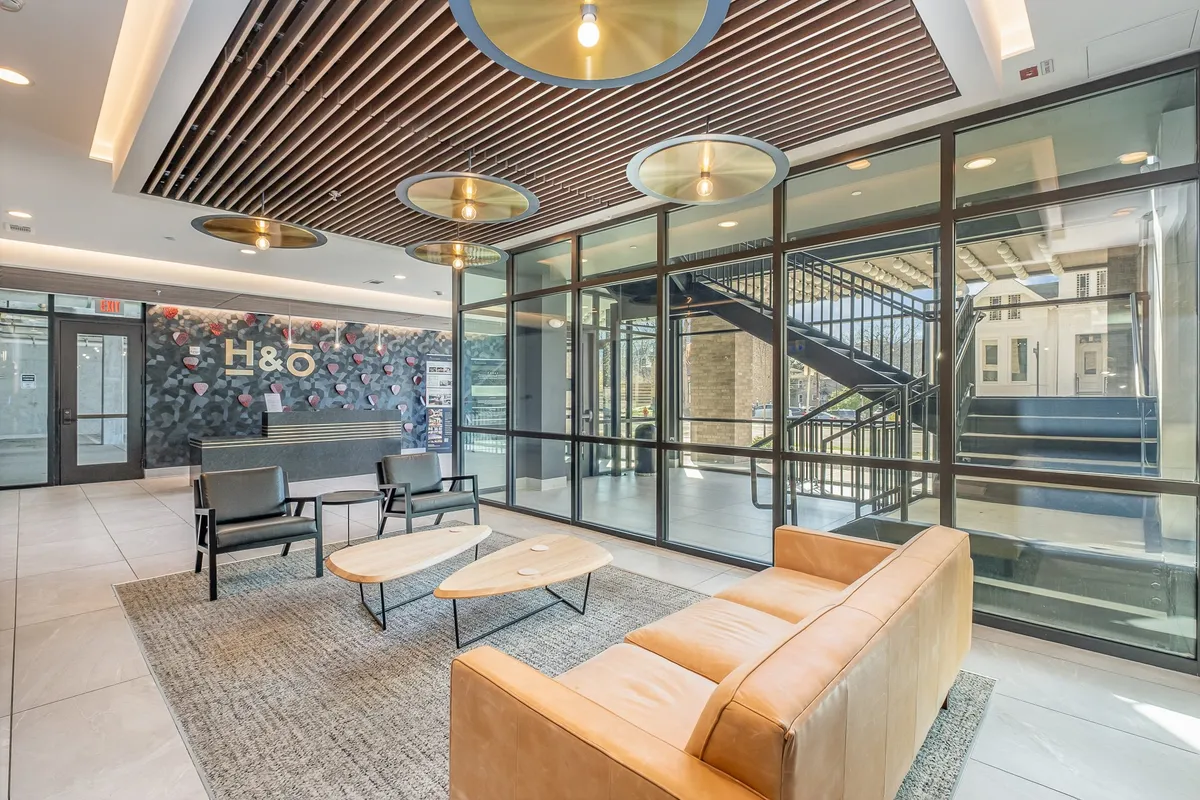The image features a contemporary entryway with a leather sofa, wooden ceiling panels, a glass wall with outdoor views, and a welcoming reception desk. The overall vibe is warm and inviting, suggesting a smooth arrival and departure experience for guests.