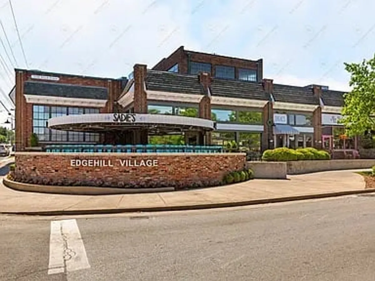 A bright daytime exterior view of Edgehill Village, featuring chic signage, the entrance to Sade's restaurant, and modern architecture. The scene captures people strolling, shopping, and enjoying the lively community atmosphere.